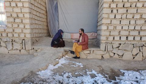 A woman sits on the ground with snow around, speaking to a man in Afghanistan