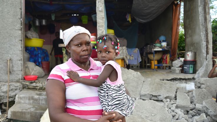 Tessa, a mother of 10, stands in front of her ruined house in Haiti holding her daughter in her arms Photo: Fabienne Douce/ActionAid.